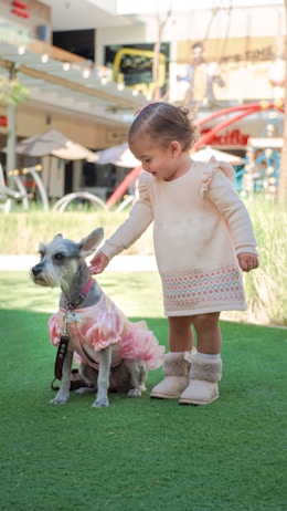 A young child in a cream-colored dress and fluffy boots is gently touching the head of a small dog dressed in a pink outfit. They are standing on a green patch of artificial grass in what appears to be a commercial outdoor area, with blurred backgrounds of benches, umbrellas, and some building structures.