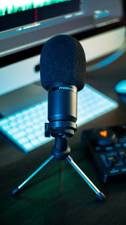 A black microphone mounted on a tripod stand sits in front of a computer screen displaying audio waveforms. The setup includes a keyboard and an audio mixer, all placed on a dark surface.