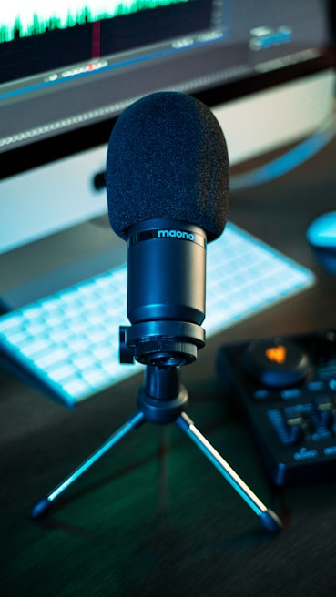 A black microphone mounted on a tripod stand sits in front of a computer screen displaying audio waveforms. The setup includes a keyboard and an audio mixer, all placed on a dark surface.