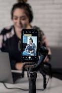 A person is seated in front of a laptop, with a smartphone attached to a gimbal in the foreground. The smartphone's screen displays a video or photo of the person, who is wearing headphones and sitting in a casual setting. The background features a white brick wall, giving a modern and professional atmosphere.