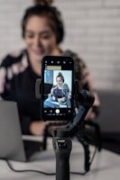 A person is seated in front of a laptop, with a smartphone attached to a gimbal in the foreground. The smartphone's screen displays a video or photo of the person, who is wearing headphones and sitting in a casual setting. The background features a white brick wall, giving a modern and professional atmosphere.