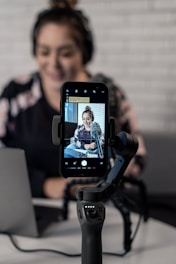 A person is seated in front of a laptop, with a smartphone attached to a gimbal in the foreground. The smartphone's screen displays a video or photo of the person, who is wearing headphones and sitting in a casual setting. The background features a white brick wall, giving a modern and professional atmosphere.