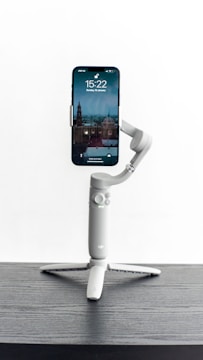A smartphone is mounted on a compact, modern-looking gimbal placed on a dark wooden surface. The phone screen displays the time, date, and a scenic background featuring architectural structures. The gimbal has a sleek, gray design with visible buttons and a sturdy tripod base.