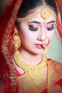 A woman adorned in traditional bridal jewelry, wearing a rich red veil. She has elaborate gold jewelry including earrings, a large necklace, and a forehead ornament. Her makeup is detailed, with bold eyeliner and red lipstick.