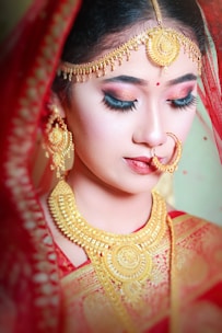 A woman adorned in traditional bridal jewelry, wearing a rich red veil. She has elaborate gold jewelry including earrings, a large necklace, and a forehead ornament. Her makeup is detailed, with bold eyeliner and red lipstick.