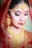 A woman adorned in traditional bridal jewelry, wearing a rich red veil. She has elaborate gold jewelry including earrings, a large necklace, and a forehead ornament. Her makeup is detailed, with bold eyeliner and red lipstick.