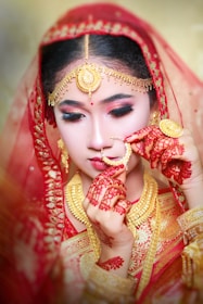 A woman clad in traditional attire with intricate gold jewelry and a red veil. Her hands are adorned with henna designs, and she delicately holds a nose ring. Her makeup features bold, dramatic eye makeup and a small red bindi on her forehead.