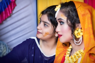 Two women in traditional attire are shown, with intricate makeup and accessories, including floral jewelry and vibrant garments. They appear to be involved in a cultural or festive event.