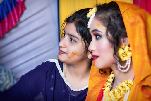 Two women in traditional attire are shown, with intricate makeup and accessories, including floral jewelry and vibrant garments. They appear to be involved in a cultural or festive event.
