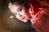 A close-up portrait of a woman wearing traditional attire, adorned with decorative floral and pearl ornaments on her hair. The woman has bright makeup, including red lipstick and eye makeup, and is wearing a red outfit.