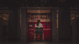 A person with long hair, seen from the back, sits on a chair in front of a vibrant red piano. The piano is inside a dimly lit room or hall, with a sign in the background reading 'La Beauté du Monde'. The atmosphere is calm and intimate.