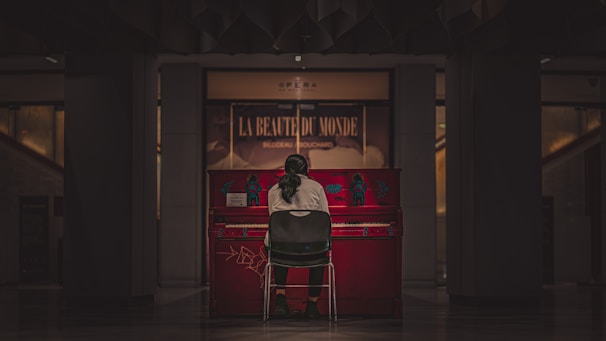 A person with long hair, seen from the back, sits on a chair in front of a vibrant red piano. The piano is inside a dimly lit room or hall, with a sign in the background reading 'La Beauté du Monde'. The atmosphere is calm and intimate.