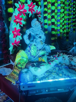 A detailed arrangement featuring a statue of Ganesha adorned with colorful garlands and surrounded by vibrant decorations and offerings. The scene includes numerous flowers and decorative cloths, contributing to a festive atmosphere. Additionally, there are various objects such as a light source and fruit placed nearby.