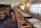 A well-decorated restaurant interior with a long row of tables set for dining, featuring patterned plates and glassware. The space has wooden chairs with cushioned seats and a long upholstered bench along the wall. Soft lighting emanates from stylish wall sconces and ceiling lights.