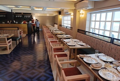 A well-decorated restaurant interior with a long row of tables set for dining, featuring patterned plates and glassware. The space has wooden chairs with cushioned seats and a long upholstered bench along the wall. Soft lighting emanates from stylish wall sconces and ceiling lights.