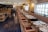 A well-decorated restaurant interior with a long row of tables set for dining, featuring patterned plates and glassware. The space has wooden chairs with cushioned seats and a long upholstered bench along the wall. Soft lighting emanates from stylish wall sconces and ceiling lights.