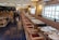 A well-decorated restaurant interior with a long row of tables set for dining, featuring patterned plates and glassware. The space has wooden chairs with cushioned seats and a long upholstered bench along the wall. Soft lighting emanates from stylish wall sconces and ceiling lights.