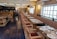 A well-decorated restaurant interior with a long row of tables set for dining, featuring patterned plates and glassware. The space has wooden chairs with cushioned seats and a long upholstered bench along the wall. Soft lighting emanates from stylish wall sconces and ceiling lights.