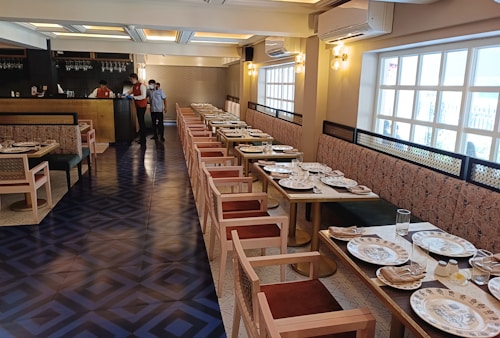 A well-decorated restaurant interior with a long row of tables set for dining, featuring patterned plates and glassware. The space has wooden chairs with cushioned seats and a long upholstered bench along the wall. Soft lighting emanates from stylish wall sconces and ceiling lights.