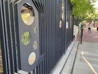 A black metal fence with circular cut-out windows is positioned at an angle, leading towards a brick-paved walkway where a few people are walking. Trees with green leaves provide shade along the pathway. Various signs are attached to the fence.