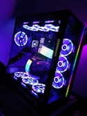 A sleek, modern desktop computer with a glass case displaying internal components illuminated by colorful RGB lighting. The setup includes multiple cooling fans arranged on the top and bottom, each glowing with vibrant light patterns. The interior showcases a prominent RGB strip and various components, all bathed in hues of blue, purple, and green.