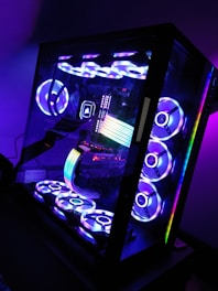 A sleek, modern desktop computer with a glass case displaying internal components illuminated by colorful RGB lighting. The setup includes multiple cooling fans arranged on the top and bottom, each glowing with vibrant light patterns. The interior showcases a prominent RGB strip and various components, all bathed in hues of blue, purple, and green.