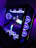 A sleek, modern desktop computer with a glass case displaying internal components illuminated by colorful RGB lighting. The setup includes multiple cooling fans arranged on the top and bottom, each glowing with vibrant light patterns. The interior showcases a prominent RGB strip and various components, all bathed in hues of blue, purple, and green.