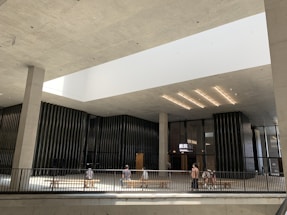 A modern indoor space with high ceilings and an open design features a cinema entrance showing glowing signage. The architecture is characterized by minimalistic, clean lines and large vertical columns. Natural light pours in from a large, rectangular skylight above. People are sitting on benches and walking around the space.