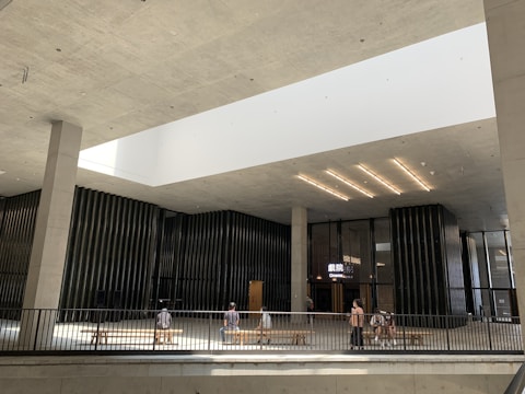 A modern indoor space with high ceilings and an open design features a cinema entrance showing glowing signage. The architecture is characterized by minimalistic, clean lines and large vertical columns. Natural light pours in from a large, rectangular skylight above. People are sitting on benches and walking around the space.