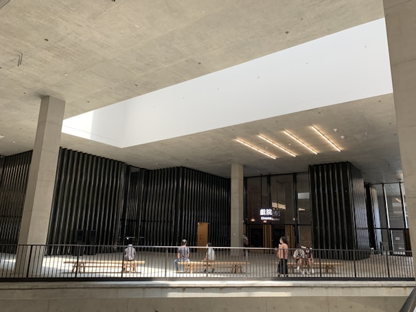 A modern indoor space with high ceilings and an open design features a cinema entrance showing glowing signage. The architecture is characterized by minimalistic, clean lines and large vertical columns. Natural light pours in from a large, rectangular skylight above. People are sitting on benches and walking around the space.