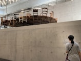 A concrete indoor space features an elevated round platform covered with a variety of chairs in different styles and colors. A person stands below, reading information on a wall-mounted sign. The room has a modern, minimalistic design with visible ceiling beams and lighting.