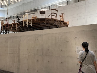 A concrete indoor space features an elevated round platform covered with a variety of chairs in different styles and colors. A person stands below, reading information on a wall-mounted sign. The room has a modern, minimalistic design with visible ceiling beams and lighting.