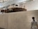 A concrete indoor space features an elevated round platform covered with a variety of chairs in different styles and colors. A person stands below, reading information on a wall-mounted sign. The room has a modern, minimalistic design with visible ceiling beams and lighting.