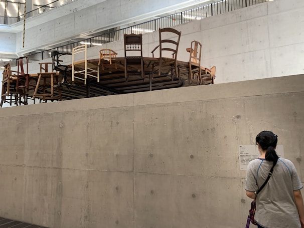 A concrete indoor space features an elevated round platform covered with a variety of chairs in different styles and colors. A person stands below, reading information on a wall-mounted sign. The room has a modern, minimalistic design with visible ceiling beams and lighting.