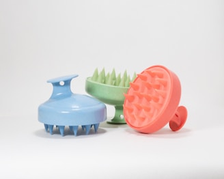 Three round scalp massagers with handle, featuring spiked bristles and made of plastic material. They are positioned side by side on a plain white background. Each massager is a different color: blue, green, and orange.