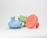Three round scalp massagers with handle, featuring spiked bristles and made of plastic material. They are positioned side by side on a plain white background. Each massager is a different color: blue, green, and orange.