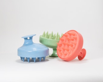Three round scalp massagers with handle, featuring spiked bristles and made of plastic material. They are positioned side by side on a plain white background. Each massager is a different color: blue, green, and orange.