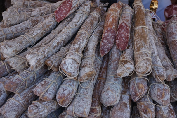 A pile of cured sausages is arranged closely together, wrapped with strings. The outer surface of the sausages has a powdery appearance, likely due to the curing process. Some sausages have been cut to reveal a marbled interior of meat and fat.