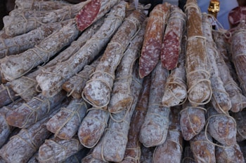 A pile of cured sausages is arranged closely together, wrapped with strings. The outer surface of the sausages has a powdery appearance, likely due to the curing process. Some sausages have been cut to reveal a marbled interior of meat and fat.