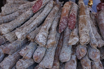 A pile of cured sausages is arranged closely together, wrapped with strings. The outer surface of the sausages has a powdery appearance, likely due to the curing process. Some sausages have been cut to reveal a marbled interior of meat and fat.