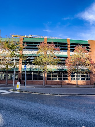 A modern multi-story car park is seen with a facade of red brick and green roofing. The structure is flanked by tall trees with autumnal foliage, showing a mix of green, yellow, and red leaves. A clear blue sky forms the backdrop, while a crane is visible at the top. A roadway runs in front of the building, with a traffic sign and painted markings on the asphalt.
