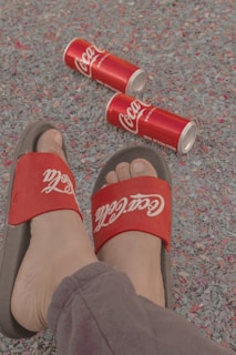 A person wearing red Coca-Cola branded sandals is standing on a textured surface covered with colorful confetti-like material. Several red Coca-Cola cans are scattered on the ground nearby.