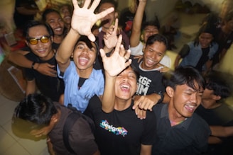 A group of young people is gathered closely together, smiling and reaching towards the camera with a sense of excitement and camaraderie. They appear to be enjoying a lively moment indoors, possibly at a party or event.