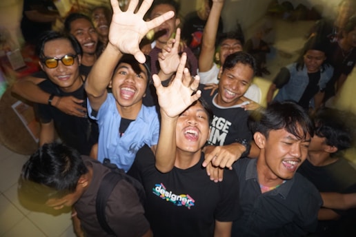 A group of young people is gathered closely together, smiling and reaching towards the camera with a sense of excitement and camaraderie. They appear to be enjoying a lively moment indoors, possibly at a party or event.