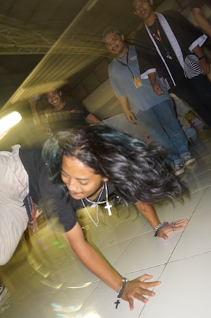 A dynamic scene with a person performing a breakdance move on a tiled floor. The individual is wearing a black shirt, beige pants, and several necklaces. Their long hair is flowing with the motion. In the background, three people are smiling and observing the performance. The lighting creates motion blur, adding a sense of movement to the image.