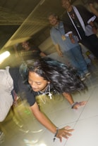 A dynamic scene with a person performing a breakdance move on a tiled floor. The individual is wearing a black shirt, beige pants, and several necklaces. Their long hair is flowing with the motion. In the background, three people are smiling and observing the performance. The lighting creates motion blur, adding a sense of movement to the image.