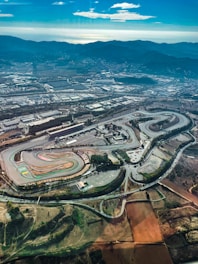 Aerial view of a racetrack with winding loops and straight sections surrounded by various buildings and infrastructure. The landscape features a mix of natural terrain, roads, and parking areas. In the distance, mountain ranges create a scenic backdrop under a partly cloudy sky.