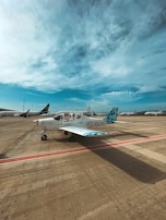 A small airplane is parked on an airport tarmac under a partly cloudy blue sky. The aircraft is white with blue and teal accents, marked with the identifiers EC-MLI and EAS Barcelona on the fuselage. Other larger airplanes are visible in the background, indicating a busy airport setting.