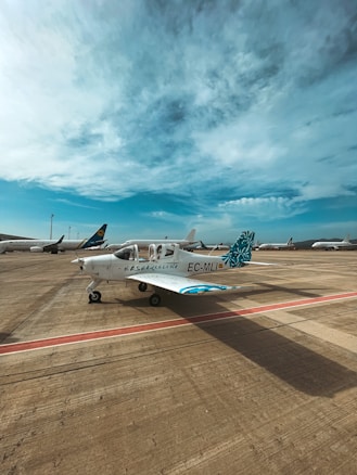 A small airplane is parked on an airport tarmac under a partly cloudy blue sky. The aircraft is white with blue and teal accents, marked with the identifiers EC-MLI and EAS Barcelona on the fuselage. Other larger airplanes are visible in the background, indicating a busy airport setting.