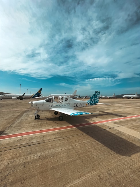 A small airplane is parked on an airport tarmac under a partly cloudy blue sky. The aircraft is white with blue and teal accents, marked with the identifiers EC-MLI and EAS Barcelona on the fuselage. Other larger airplanes are visible in the background, indicating a busy airport setting.